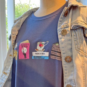 May include: A navy blue t-shirt with a graphic design featuring three book covers. The book covers feature the titles "Modesty Blaise", "Funeral in Berlin", and "Spook's Deadliest", with illustrations of a woman, a teacup, and a skull.
