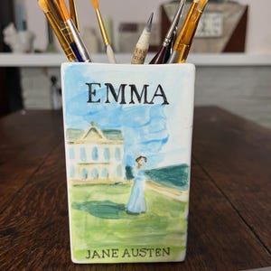 Pode incluir: Um porta-lápis de cerâmica branca com uma pintura em aquarela de uma cena de "Emma" de Jane Austen. O porta-lápis exibe o título "EMMA" e o nome do autor. Vários pincéis estão dentro do porta-lápis, e um está sobre a superfície de madeira.