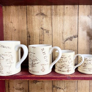 May include: A set of four cream-coloured ceramic mugs of varying sizes, each featuring a brown printed map of Middle-earth. The mugs display geographical labels such as Forodwaith, Rhun, Mordor, Khand, and Near Harad, with a curved handle on each.