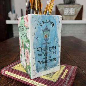 May include: A ceramic pencil holder featuring illustrations from "The Secret Garden" and "The Lion, the Witch, and the Wardrobe" by C.S. Lewis. The holder is filled with paintbrushes and rests on a copy of "The Wind in the Willows" by Kenneth Grahame.