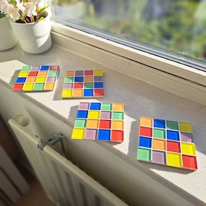 May include: Four colorful square coasters made of mosaic tiles. Each coaster has a different color scheme with shades of yellow, green, blue, red, and purple.