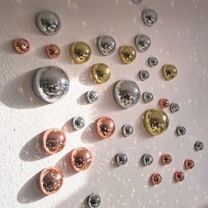 May include: A collection of decorative disco balls in various sizes and colors, including silver, gold, and copper. The balls are arranged in a random pattern on a white wall.