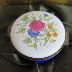 Peut inclure: Une petite boîte à bijoux ronde en porcelaine blanche avec une base en émail bleu. Le couvercle présente un motif floral peint à la main avec une rose rose, des baies bleues et des fleurs jaunes.