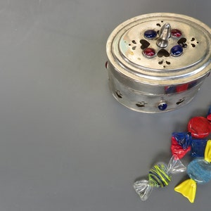 Può includere: Una scatola ovale d'argento con un coperchio decorativo e accenti in vetro colorato. La scatola è aperta e ci sono diversi pezzi di caramelle di vetro colorato sparsi intorno.