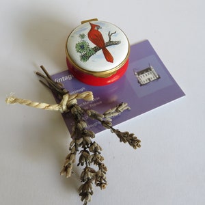 Peut inclure: Une boîte à bijoux en porcelaine rouge et blanche avec un couvercle doré. Le couvercle présente une image peinte d'un cardinal perché sur une branche. La boîte est posée sur une carte violette avec du texte blanc.