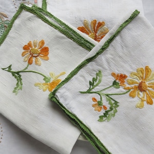 May include: Two white linen napkins with green embroidered borders and yellow and orange floral designs.