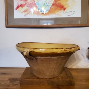 Puede incluir: Un cuenco grande y antiguo de cerámica de color marrón claro con un borde ancho y ligeramente acampanado y una base redondeada. El cuenco se asienta sobre una base cuadrada de madera. Una pintura de acuarela enmarcada está en el fondo.