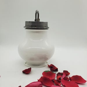 Pode incluir: Jarro de cerâmica branca com tampa prateada e alça de metal. O jarro está sobre uma superfície branca com pétalos de rosa vermelhos espalhados ao redor.