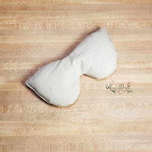 Pu&ograve; includere: Un cuscino bianco per gli occhi, sagomato per adattarsi al viso, &egrave; appoggiato su una superficie di legno marrone chiaro. Il cuscino &egrave; realizzato in un tessuto morbido e di colore chiaro. Il design &egrave; semplice e funzionale, suggerendo un focus sul relax e sul comfort.