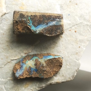 Könnte beinhalten: Zwei raue, rechteckige Stücke Boulder-Opal mit einer texturierten, erdbraunen Eisensteinmatrix. Jeder Stein zeigt eine lebendige, schillernde Ader aus blauem und grünem Opal, die horizontal über die Oberfläche des Gesteins verläuft.