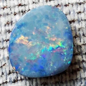 May include: A polished, oval-shaped opal gemstone. The opal has a light blue base color with iridescent flashes of yellow, green, and blue. The stone is smooth and rounded, with a subtle, internal play of color.