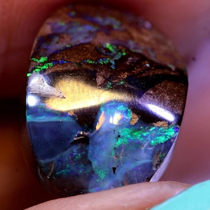 Könnte beinhalten: Ein polierter Opal-Edelstein mit einer Mischung aus Farben. Der Stein hat eine dunkelbraune Matrix mit grünen, blauen und goldenen Blitzen. Der Opal ist oval geformt und hat einen hohen Glanz, der das Licht reflektiert.