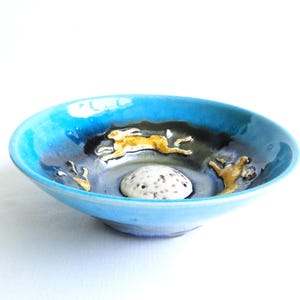 May include: A shallow ceramic bowl featuring a vibrant turquoise blue rim that transitions into a dark, moody interior. The centre contains a textured, speckled white circular element, surrounded by two golden-brown hares in motion.