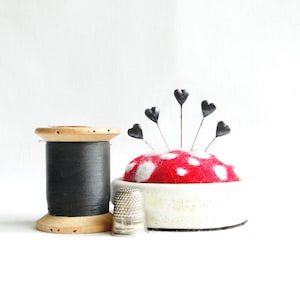 May include: A collection of sewing notions, including a wooden spool of black thread, a thimble, and a red and white pin cushion with heart-shaped pins. The items are arranged on a white background.