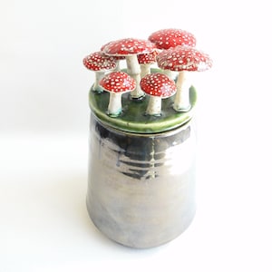 May include: A ceramic jar with a lid decorated with a cluster of red and white mushrooms. The jar has a metallic glaze and a green base.
