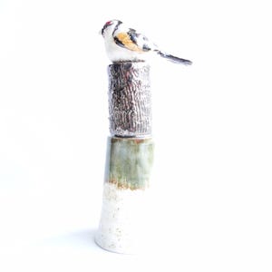 Puede incluir: Escultura de cerámica de un pájaro posado sobre una columna texturizada. El pájaro tiene marcas blancas, negras y amarillas con una mancha roja en la cabeza. La columna está compuesta por una sección texturizada plateada sobre una base verde y blanca.