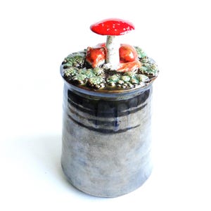 May include: A cylindrical ceramic jar with a dark, mottled grey glaze. The lid features a sculpted scene with a small orange fox curled up next to a white-stemmed red mushroom, surrounded by textured green moss-like details on the surface.