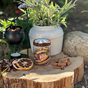 Può includere: Un'esposizione rustica con un calderone nero, un barattolo di vetro con frutta secca e un vaso di ceramica bianca con piante. L'allestimento comprende fette d'arancia essiccate, bastoncini di cannella e una candela.