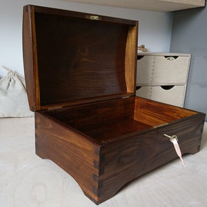 Puede incluir: Un cofre de madera rectangular con acabado marrón oscuro y tapa curva con bisagras. La caja presenta juntas de dedos visibles en las esquinas, un pestillo de metal dorado y una pequeña llave dorada insertada en la cerradura con una cinta rosa.