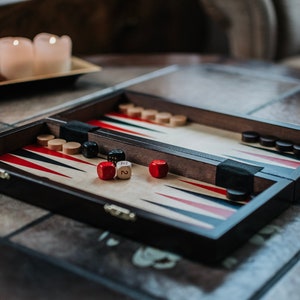Könnte beinhalten: Ein Backgammon-Spielbrett aus Holz mit schwarzen und weißen Spielsteinen, roten und weißen Würfeln und einer schwarz-weißen Spielfläche. Das Brett ist geschlossen und steht auf einer gefliesten Oberfläche.