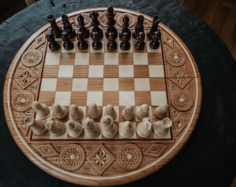 May include: A round, wooden chess set with intricately carved border details. The chessboard features a light and dark square pattern, with chess pieces in place, ready to play. The pieces are dark brown and light brown.