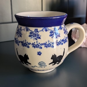 Może przedstawiać: Ceramiczny kubek o zaokrąglonym kształcie, z białą podstawą ozdobioną niebieskimi wzorami kwiatowymi i czarnymi sylwetkami kotów. Obręcz i uchwyt są w kolorze granatowym. Przedstawiono również niebieską kulkę wełny.