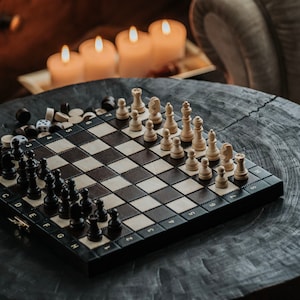 May include: A black and white chessboard with chess pieces set up on a wooden table. The chessboard is in the foreground, and there are candles in the background.