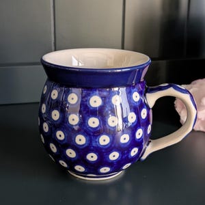 May include: A blue ceramic mug with a white interior and handle. The mug is decorated with a pattern of white dots. The mug has a rounded shape and a wide opening. The handle is a curved shape.