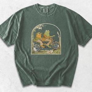 May include: A moss green short-sleeve t-shirt featuring a graphic print of two frogs riding a tandem bicycle. The illustration is framed within an arch and depicts the frogs wearing tan jackets and hats, surrounded by stylised foliage and flowers.