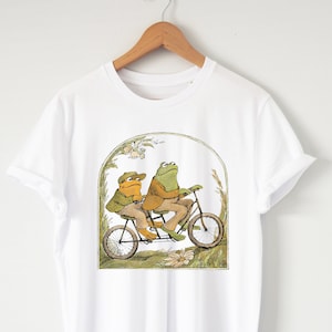 May include: White t-shirt featuring a whimsical illustration of two frogs riding a tandem bicycle. The frogs are dressed in vintage clothing, set against a backdrop of flowers and greenery. A charming and nostalgic design.
