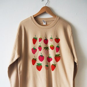 May include: A beige sweatshirt with a pattern of red and green strawberries printed on the front.