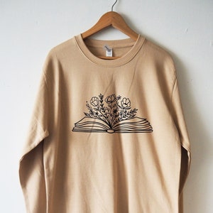 May include: A tan sweatshirt with a black graphic of an open book with flowers growing out of it.