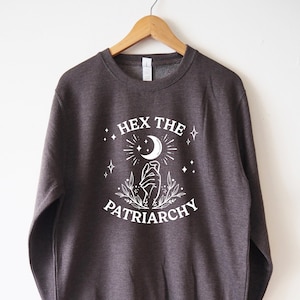 Può includere: Una felpa grigia scura con un design grafico bianco che dice "Hex the Patriarchy" con una mano che tiene una luna crescente e stelle.