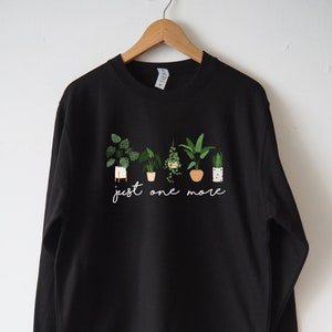 Puede incluir: Sudadera negra con un gr&aacute;fico de plantas en maceta y el texto "just one more".