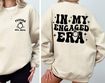 May include: A beige sweatshirt with a black graphic on the front that says "Future Mrs. Smith" with a ring design. The back of the sweatshirt has a black graphic that says "In My Engaged Era" with a ring design.
