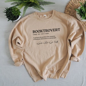 May include: Tan sweatshirt with the text "BOOKTROVERT (book-tro-vert) noun A person who prefers the company of fictional characters to real people." and a graphic of five books.