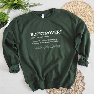 May include: A dark green sweatshirt with the text "BOOKTROVERT (book-tro-vert). noun A person who prefers the company of fictional characters to real people." printed in white. The text is surrounded by five small illustrations of open books.
