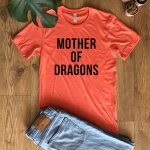 May include: Coral-colored t-shirt with the text "MOTHER OF DRAGONS" in black block letters. The shirt is paired with light blue denim jeans. The shirt is made of a soft material.