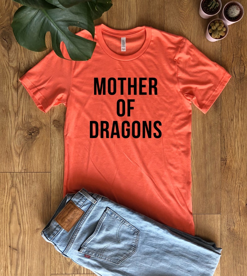 May include: Coral-colored t-shirt with the text "MOTHER OF DRAGONS" in black block letters. The shirt is paired with light blue denim jeans. The shirt is made of a soft material.