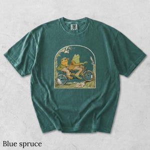 May include: A blue spruce colored short-sleeve t-shirt featuring a graphic print of two frogs riding a bicycle together. The illustration is framed within an arch and includes floral details. The shirt has a crew neckline and a visible brand tag.