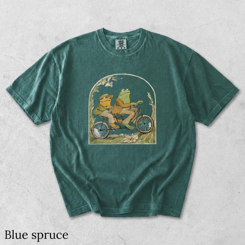 May include: A blue spruce colored short-sleeve t-shirt featuring a graphic print of two frogs riding a bicycle together. The illustration is framed within an arch and includes floral details. The shirt has a crew neckline and a visible brand tag.