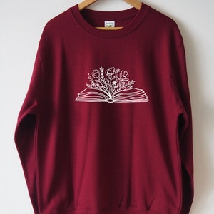 May include: A maroon sweatshirt with a white graphic of an open book with flowers growing out of it.