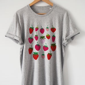 Puede incluir: Una camiseta gris con un estampado de fresas rojas y rosas en la parte delantera.