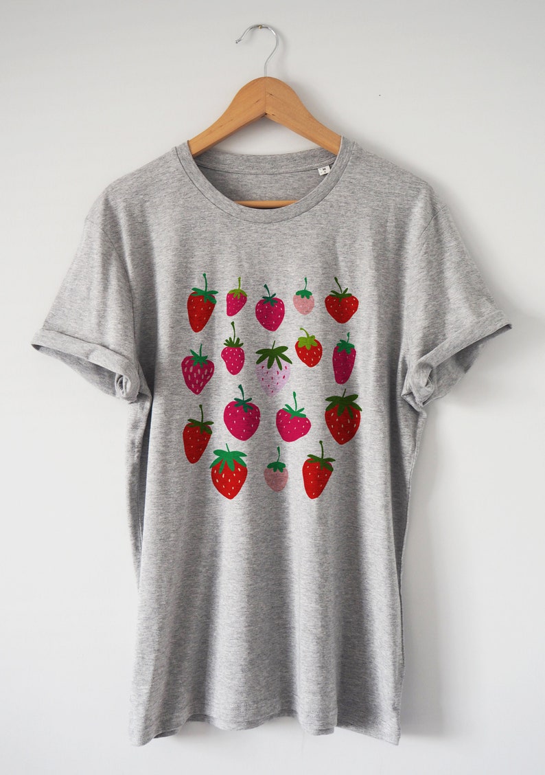 Puede incluir: Una camiseta gris con un estampado de fresas rojas y rosas en la parte delantera.