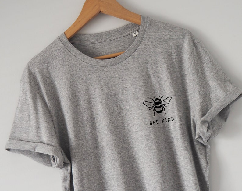 Może przedstawiać: Szary t-shirt z czarnym nadrukiem pszczoły i tekstem "BEE KIND" z przodu.