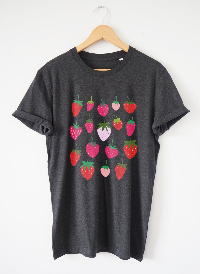 Puede incluir: Una camiseta gris oscuro con un patr&oacute;n de fresas rojas, rosas y verdes. Las fresas est&aacute;n dispuestas en un patr&oacute;n de cuadr&iacute;cula en la parte delantera de la camiseta.