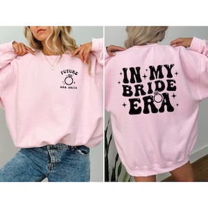 May include: Pink sweatshirt with a black graphic on the front that says "Future Mrs. Smith" with a ring and a black graphic on the back that says "In My Bride Era" with a ring.