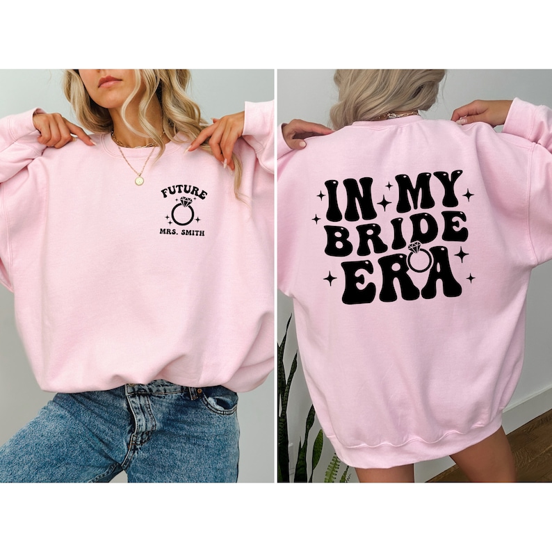 May include: Pink sweatshirt with a black graphic on the front that says "Future Mrs. Smith" with a ring and a black graphic on the back that says "In My Bride Era" with a ring.