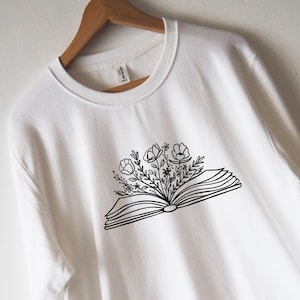 May include: White sweatshirt with a black line drawing of an open book with flowers growing out of it.