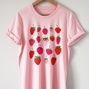 Puede incluir: Una camiseta rosa con un estampado gr&aacute;fico de fresas rojas, rosas y verdes.